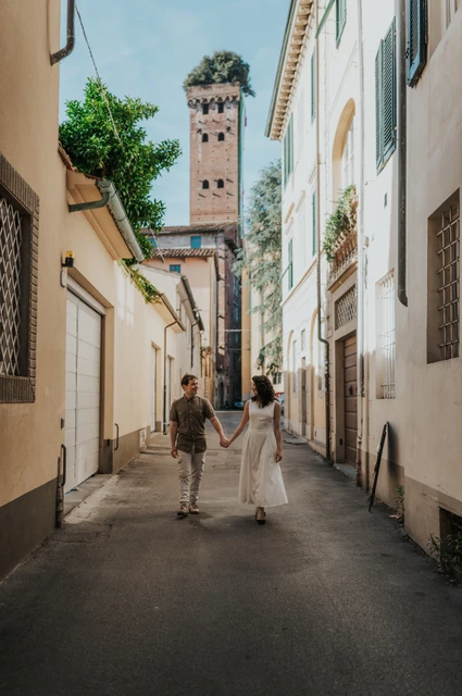 Lucca couple photoshoot: a narrow street with Guinigi Tower beyond The couple walking along a narrow old-town street with Guinigi Tower and its rooftop trees behind them during a couple photoshoot in Lucca.