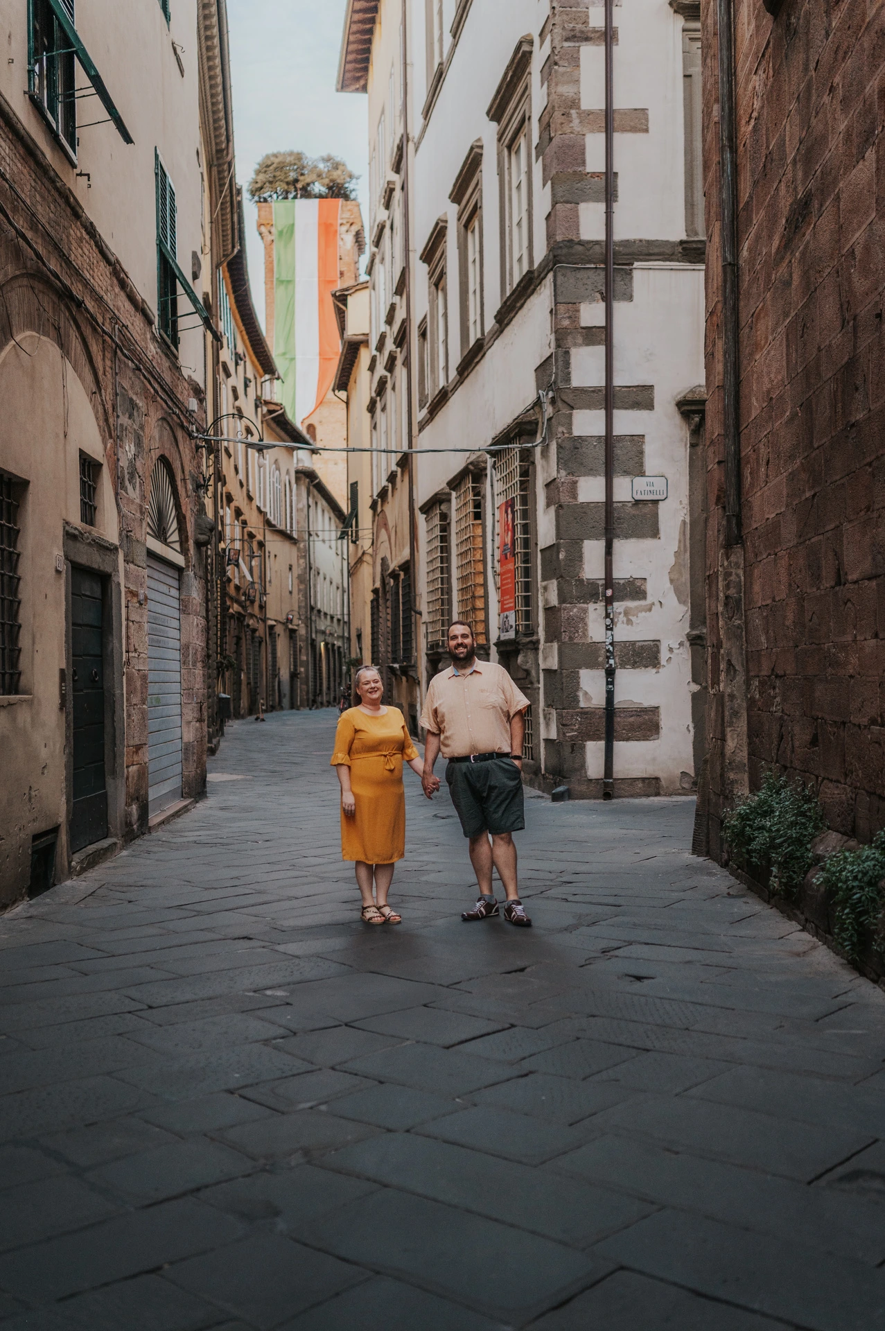 The couple walking along a narrow old-town street with Guinigi Tower and its rooftop trees in the distance during a couple photoshoot in Lucca.