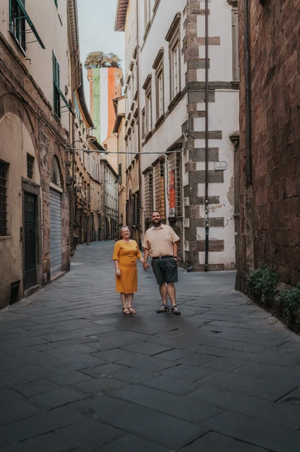 Lucca couple photoshoot: Guinigi Tower at the end of an old-town street The couple walking along a narrow old-town street with Guinigi Tower and its rooftop trees in the distance during a couple photoshoot in Lucca.