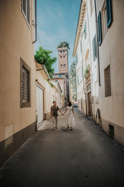 Lucca couple photoshoot: walking towards Guinigi Tower The couple walking away hand in hand along a narrow old-town street towards Guinigi Tower during a couple photoshoot in Lucca.