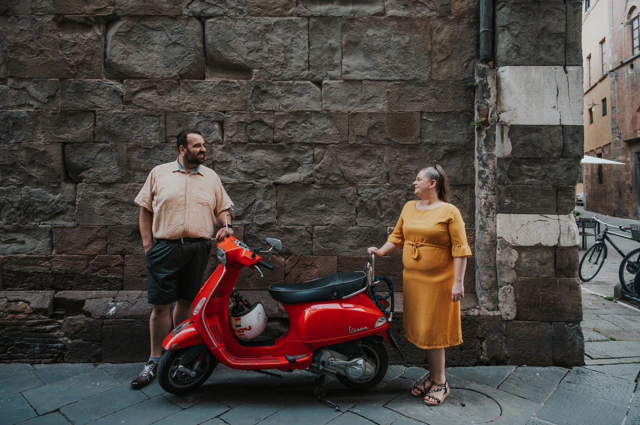Lucca couple photoshoot: a red Vespa beside the old-town stonework The couple standing on either side of a red Vespa against a textured stone wall in the old town during a couple photoshoot in Lucca.
