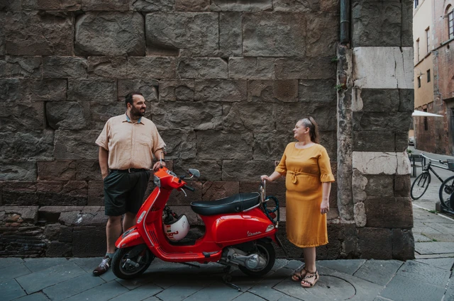 The couple standing on either side of a red Vespa against a textured stone wall in the old town during a couple photoshoot in Lucca.
