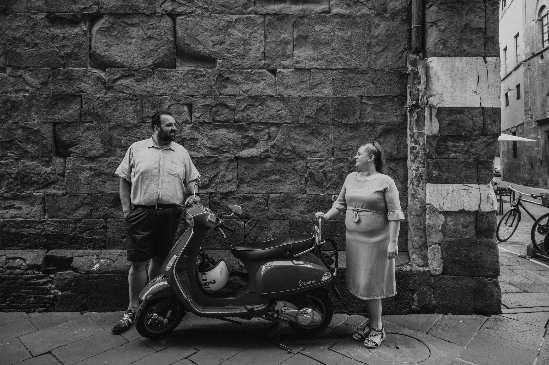 Black-and-white photograph of the couple standing apart beside a Vespa parked against a textured stone wall in the old town during a couple photoshoot in Lucca.
