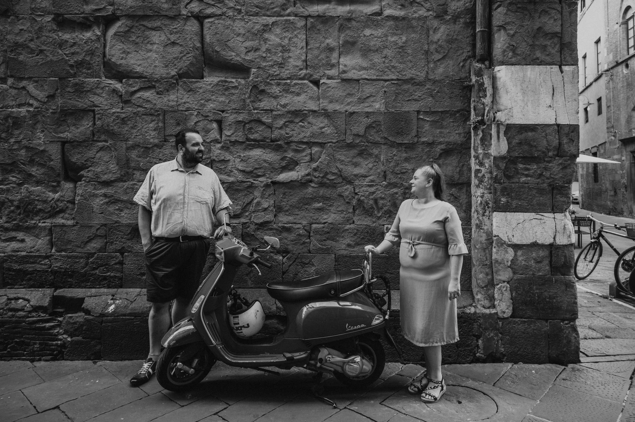 Lucca couple photoshoot: a Vespa beside a textured stone wall in black and white Black-and-white photograph of the couple standing apart beside a Vespa parked against a textured stone wall in the old town during a couple photoshoot in Lucca.