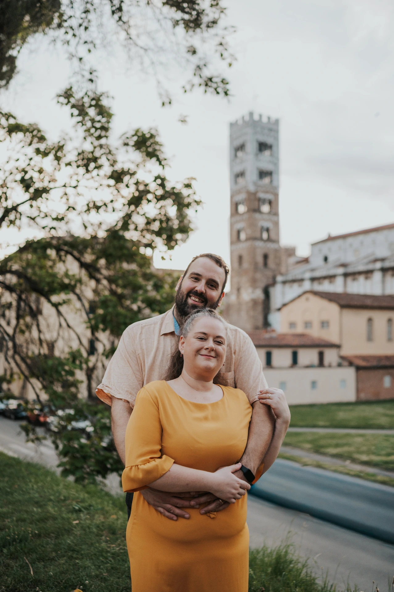Lucca couple photoshoot: a portrait with San Frediano’s bell tower The couple smiling together beneath leafy branches with San Frediano’s bell tower behind them during a couple photoshoot in Lucca.