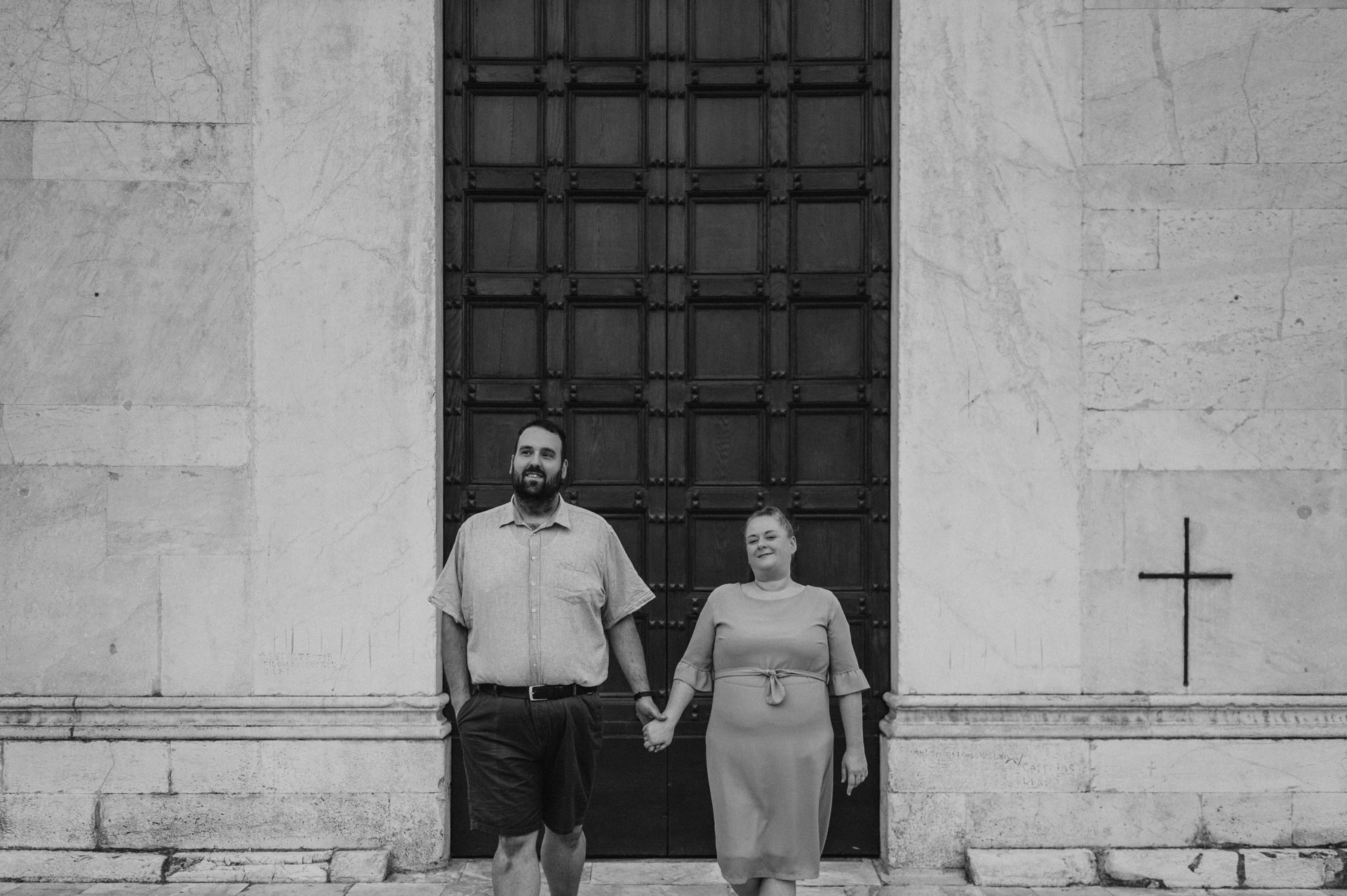 Black-and-white photograph of a closer portrait of the couple holding hands against the pale stone and wooden doors of the Basilica di San Frediano during a couple photoshoot in Lucca.