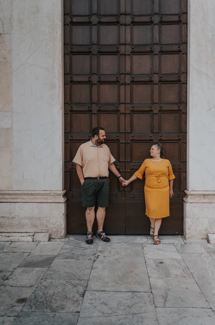 Lucca couple photoshoot: holding hands at San Frediano’s wooden doors The couple holding hands in front of the Basilica di San Frediano’s large wooden doors during a couple photoshoot in Lucca.