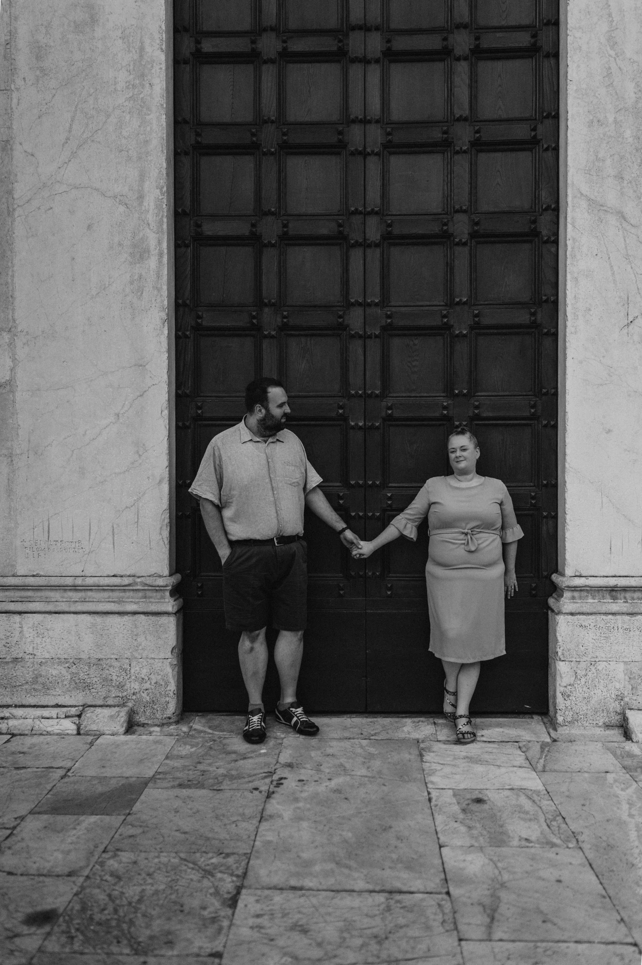 Black-and-white photograph of the couple holding hands on the stone steps in front of the Basilica di San Frediano’s wooden doors during a couple photoshoot in Lucca.