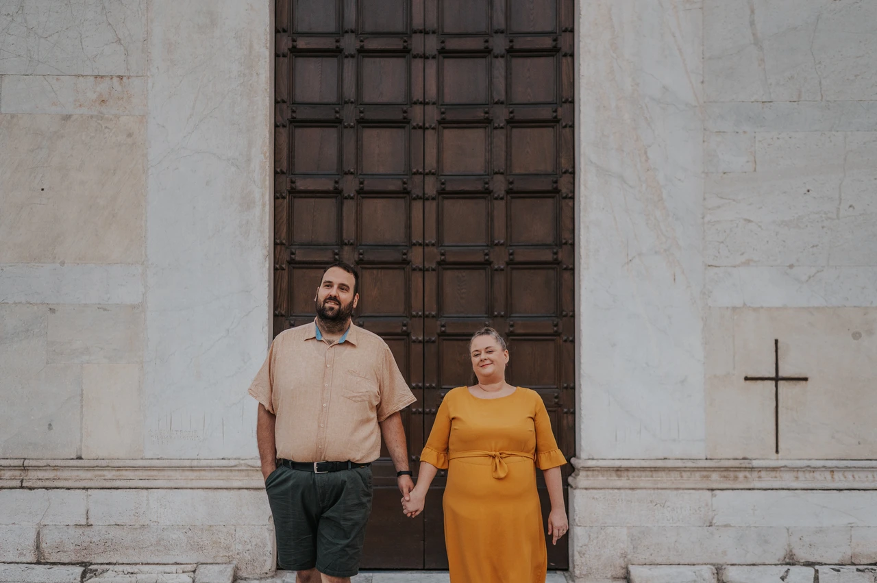 Lucca couple photoshoot: smiles beside San Frediano’s doors The couple smiling together and holding hands beside the Basilica di San Frediano’s wooden doors during a couple photoshoot in Lucca.