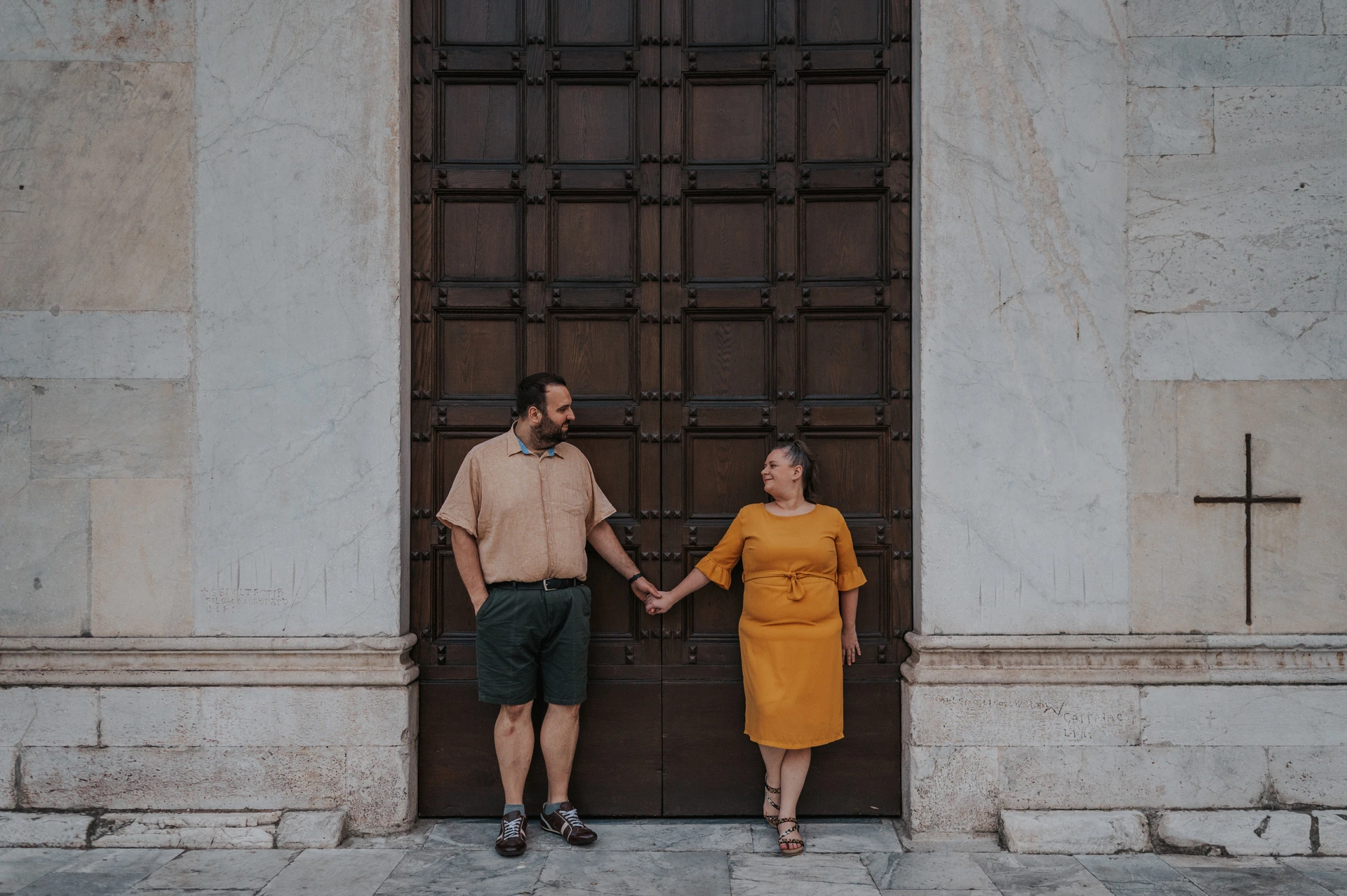 A wide portrait of the couple standing apart and holding hands on the steps of the Basilica di San Frediano during a couple photoshoot in Lucca.