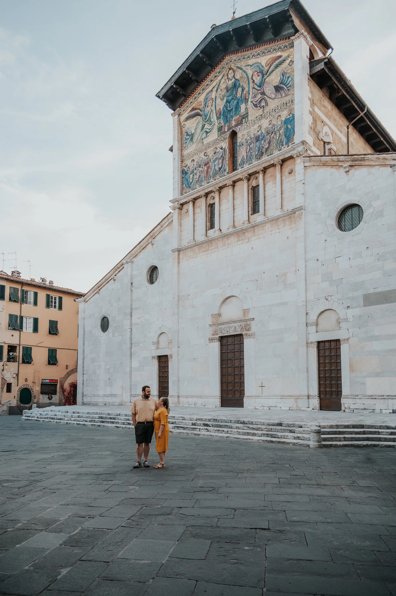 The couple standing in the square in front of the Basilica di San Frediano’s pale façade and colourful mosaic during a couple photoshoot in Lucca.