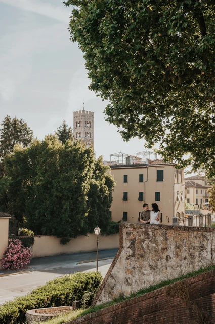 Lucca couple photoshoot: a leafy view towards San Frediano’s tower A wide view of the couple beside a stone terrace with trees and San Frediano’s bell tower beyond during a couple photoshoot in Lucca.