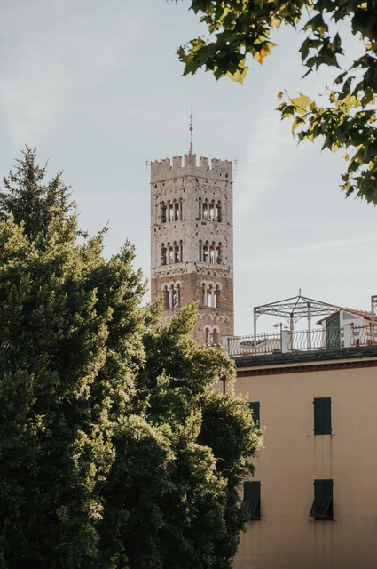 Lucca couple photoshoot: a detail of San Frediano’s bell tower San Frediano’s stone bell tower rising above leafy trees and pale buildings during a couple photoshoot in Lucca.