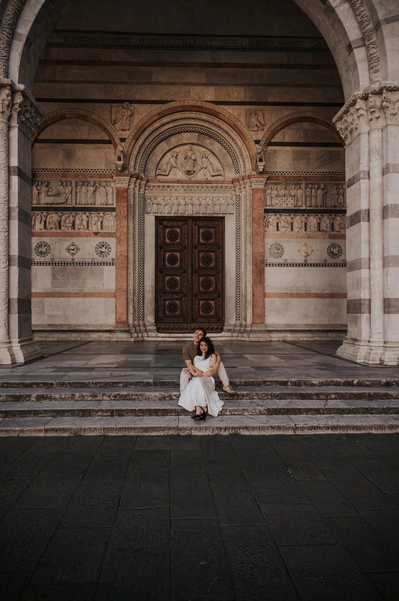 A wide view of the couple sitting on the steps beneath the Duomo di San Martino’s arched porch during a couple photoshoot in Lucca.