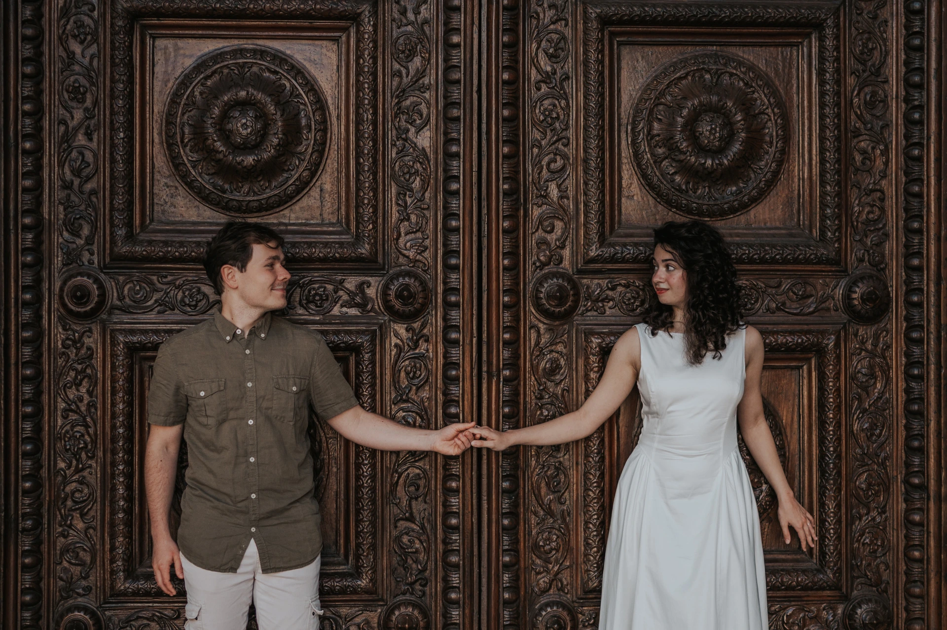 The couple looking towards each other while holding hands in front of the Duomo di San Martino’s ornate wooden doors during a couple photoshoot in Lucca.