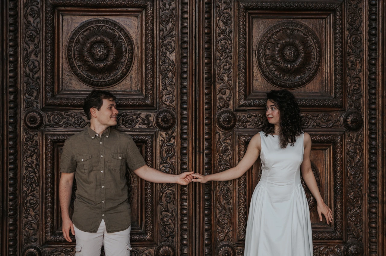 Lucca couple photoshoot: a closer portrait at the carved Duomo doors The couple looking towards each other while holding hands in front of the Duomo di San Martino’s ornate wooden doors during a couple photoshoot in Lucca.