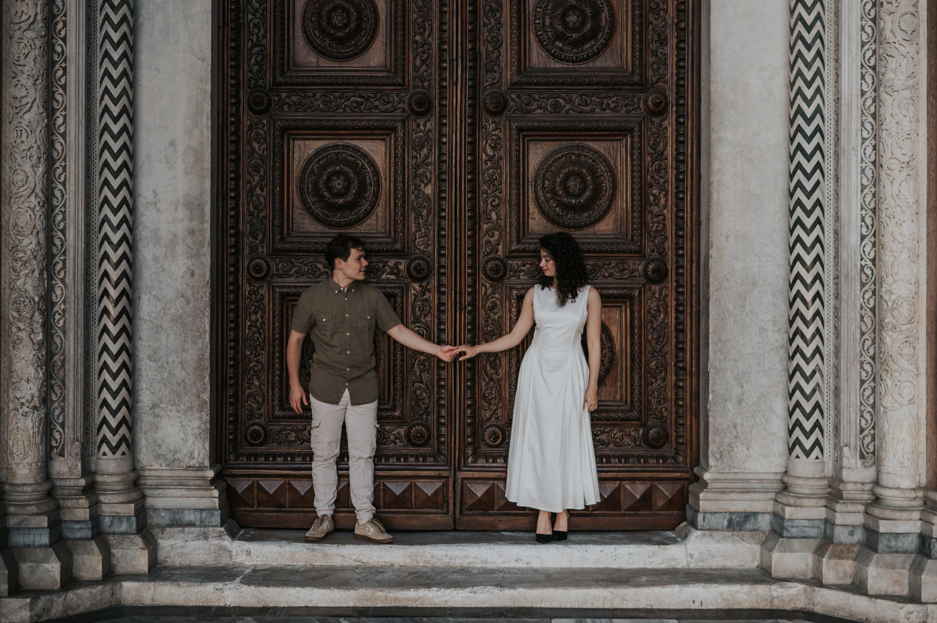 Lucca couple photoshoot: holding hands at the Duomo’s wooden doors A wide portrait of the couple standing apart and holding hands in front of the Duomo di San Martino’s carved doors during a couple photoshoot in Lucca.