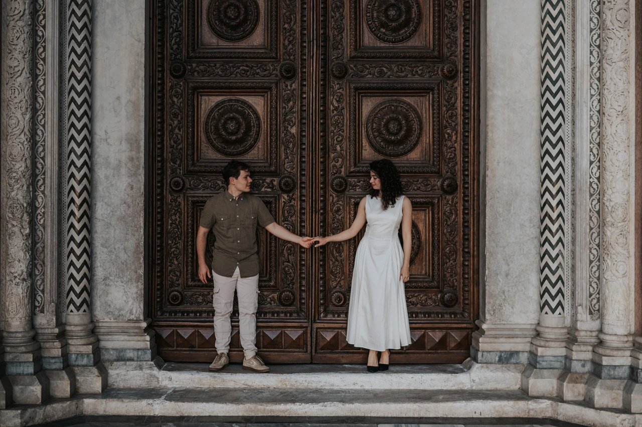 Lucca couple photoshoot: holding hands at the Duomo’s wooden doors A wide portrait of the couple standing apart and holding hands in front of the Duomo di San Martino’s carved doors during a couple photoshoot in Lucca.