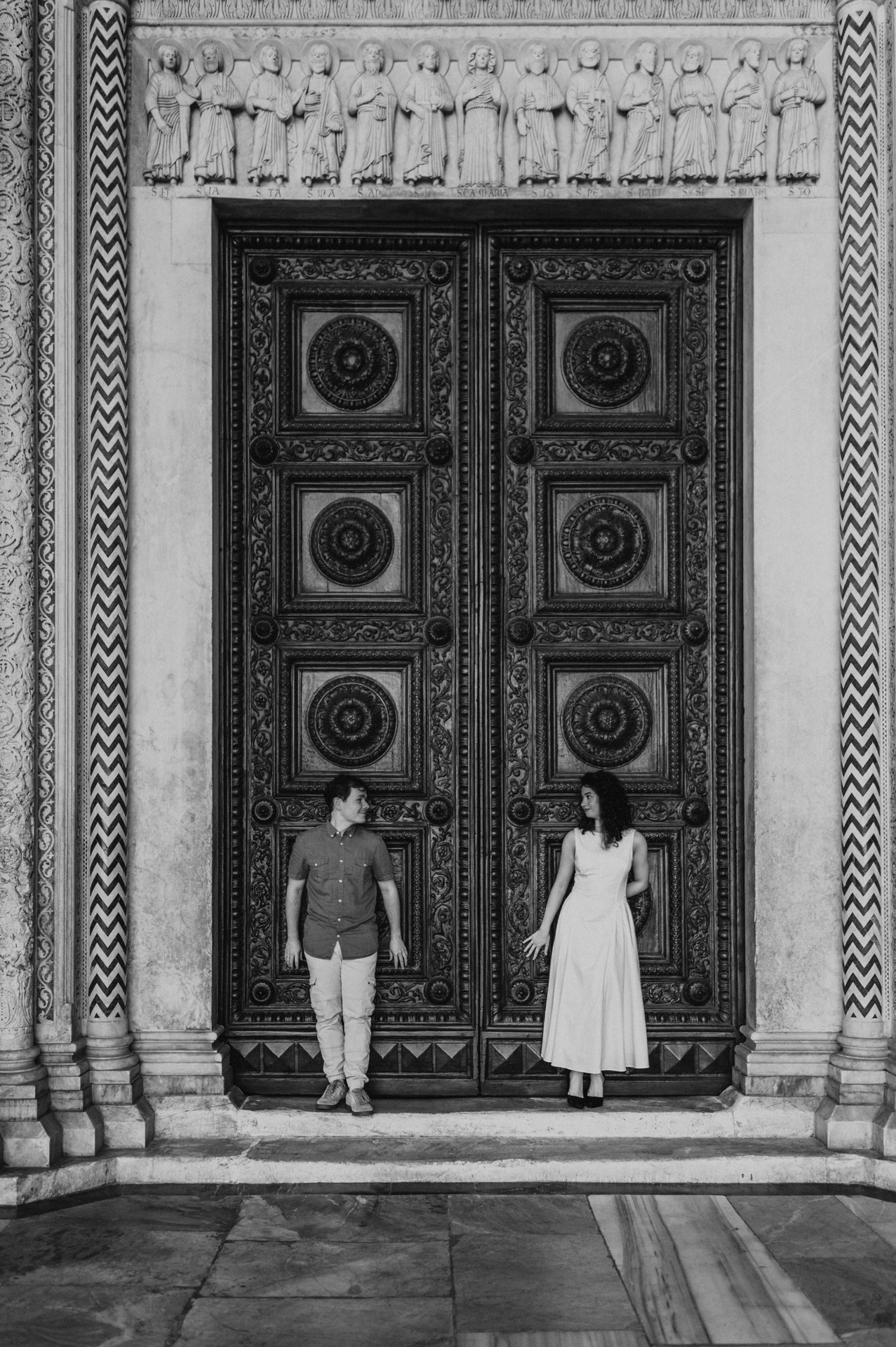 Black-and-white photograph of the couple standing apart on the steps in front of the Duomo di San Martino’s tall wooden doors during a couple photoshoot in Lucca.