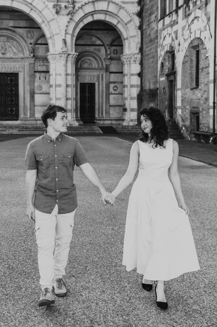 Lucca couple photoshoot: a walk together outside the Duomo in black and white Black-and-white photograph of the couple walking hand in hand across the square outside the Duomo di San Martino during a couple photoshoot in Lucca.