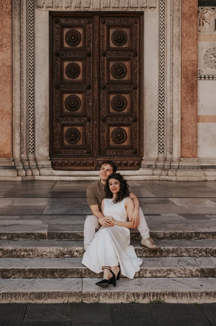 Lucca couple photoshoot: a seated portrait on San Martino’s steps The couple sitting close together on the stone steps with the Duomo di San Martino’s carved doors behind them during a couple photoshoot in Lucca.