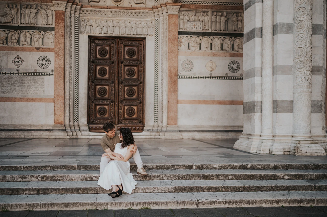 Lucca couple photoshoot: a wide view of the Duomo’s stone steps A wide portrait of the couple sitting together on the steps below the Duomo di San Martino’s carved stonework during a couple photoshoot in Lucca.