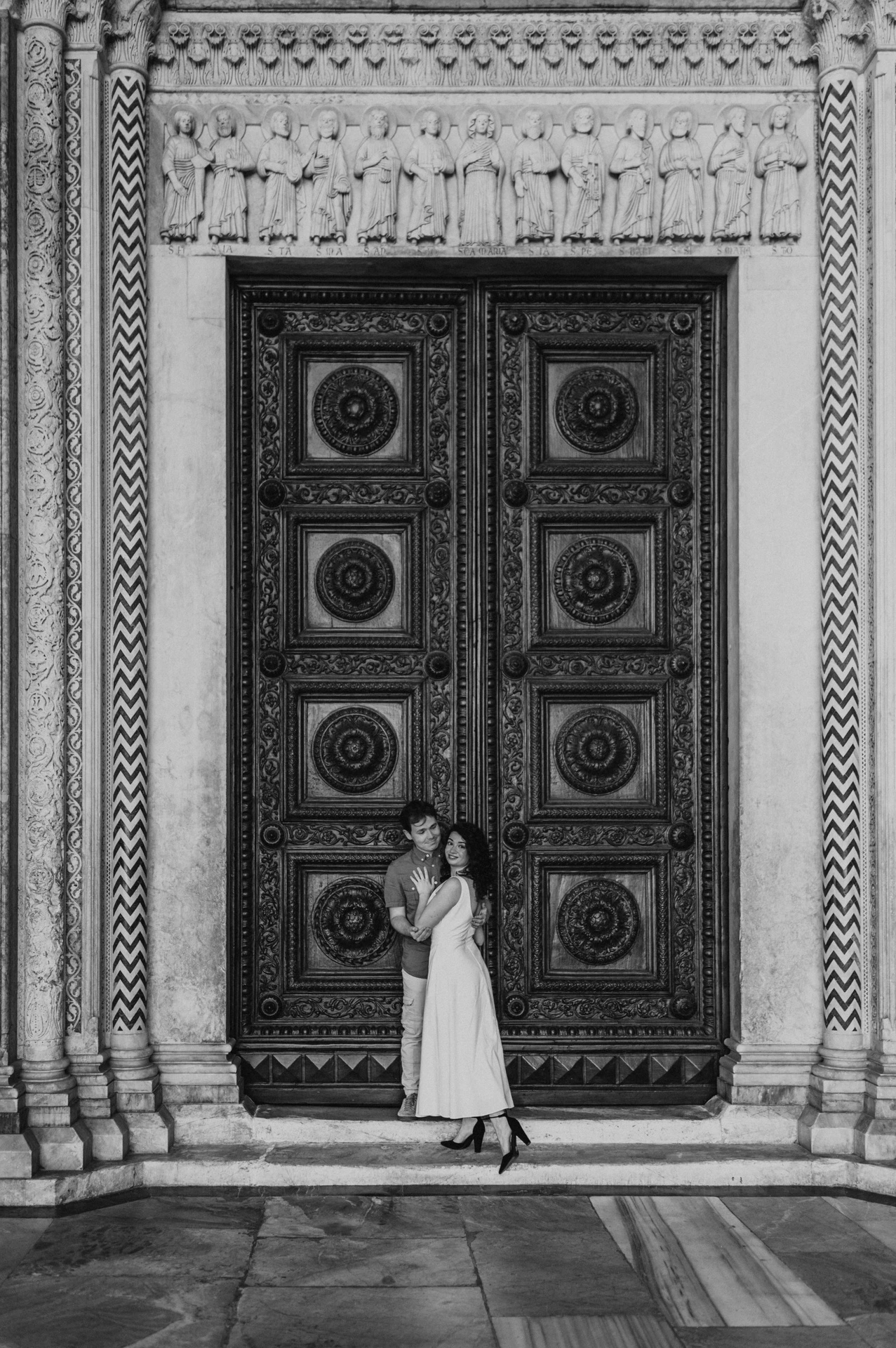 Black-and-white photograph of the couple standing close beneath the patterned stone surround of the Duomo di San Martino’s wooden doors during a couple photoshoot in Lucca.