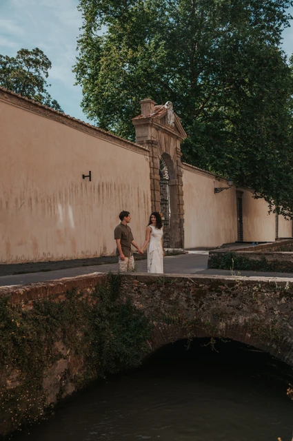 Lucca couple photoshoot: a portrait on the Via del Fosso bridge The couple standing together on the stone bridge on Via del Fosso beside a pale wall and leafy trees during a couple photoshoot in Lucca.