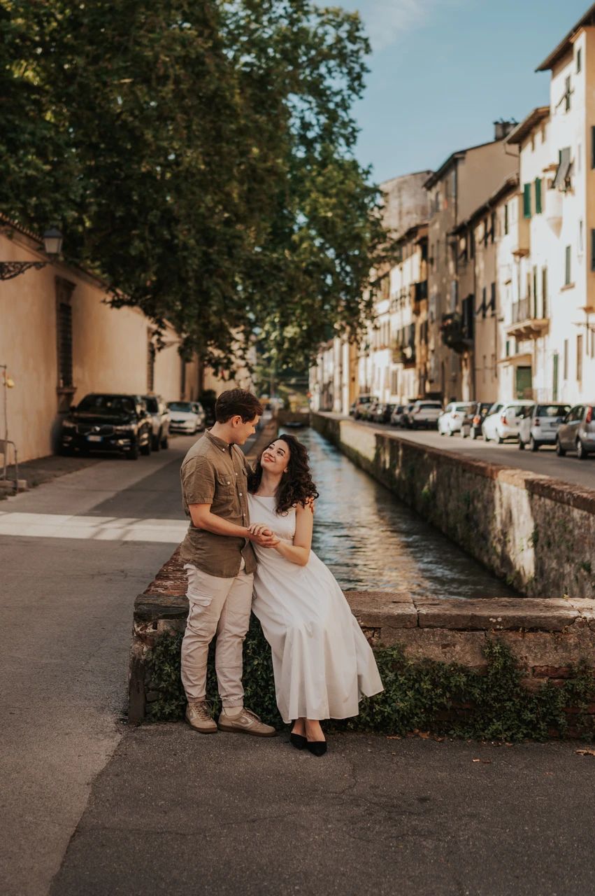 Lucca couple photoshoot: a seated portrait on Via del Fosso The couple sitting close on a stone parapet on Via del Fosso beneath leafy trees during a couple photoshoot in Lucca.