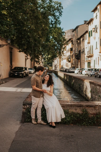 Lucca couple photoshoot: a seated portrait on Via del Fosso The couple sitting close on a stone parapet on Via del Fosso beneath leafy trees during a couple photoshoot in Lucca.