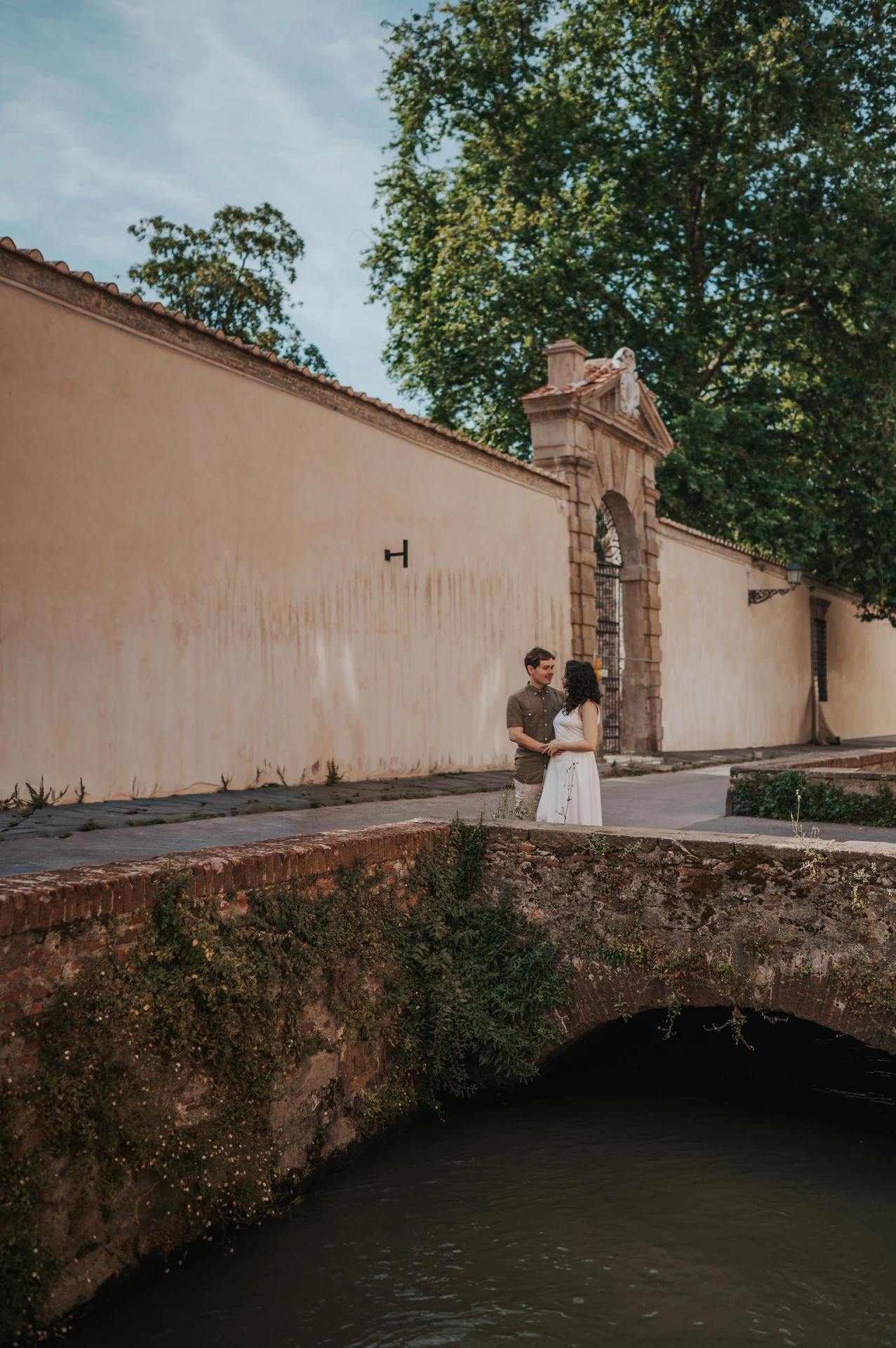 The couple standing close on a small stone bridge on Via del Fosso, with a pale wall and trees behind them during a couple photoshoot in Lucca.
