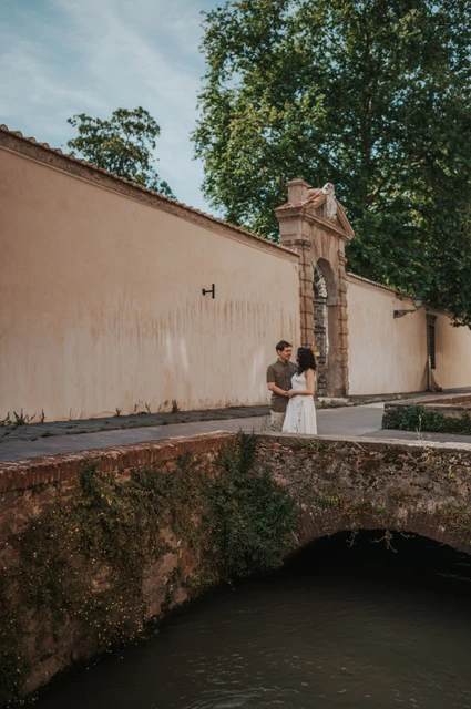 Lucca couple photoshoot: a stone bridge on Via del Fosso The couple standing close on a small stone bridge on Via del Fosso, with a pale wall and trees behind them during a couple photoshoot in Lucca.