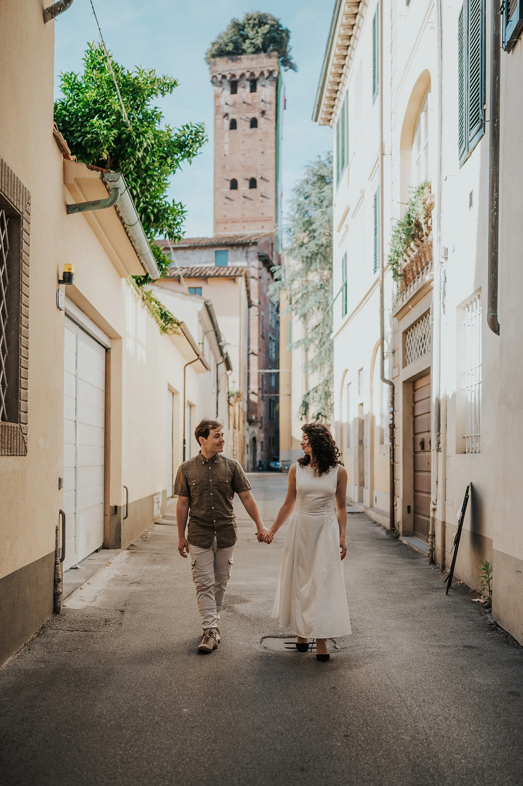 Lucca photographer captures a couple walking along a narrow lane with a tower visible at the end.