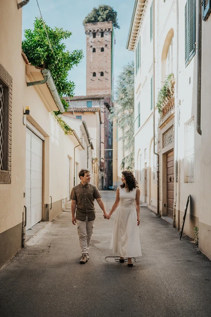 Lucca photographer captures a couple walking along a narrow lane with a tower visible at the end.