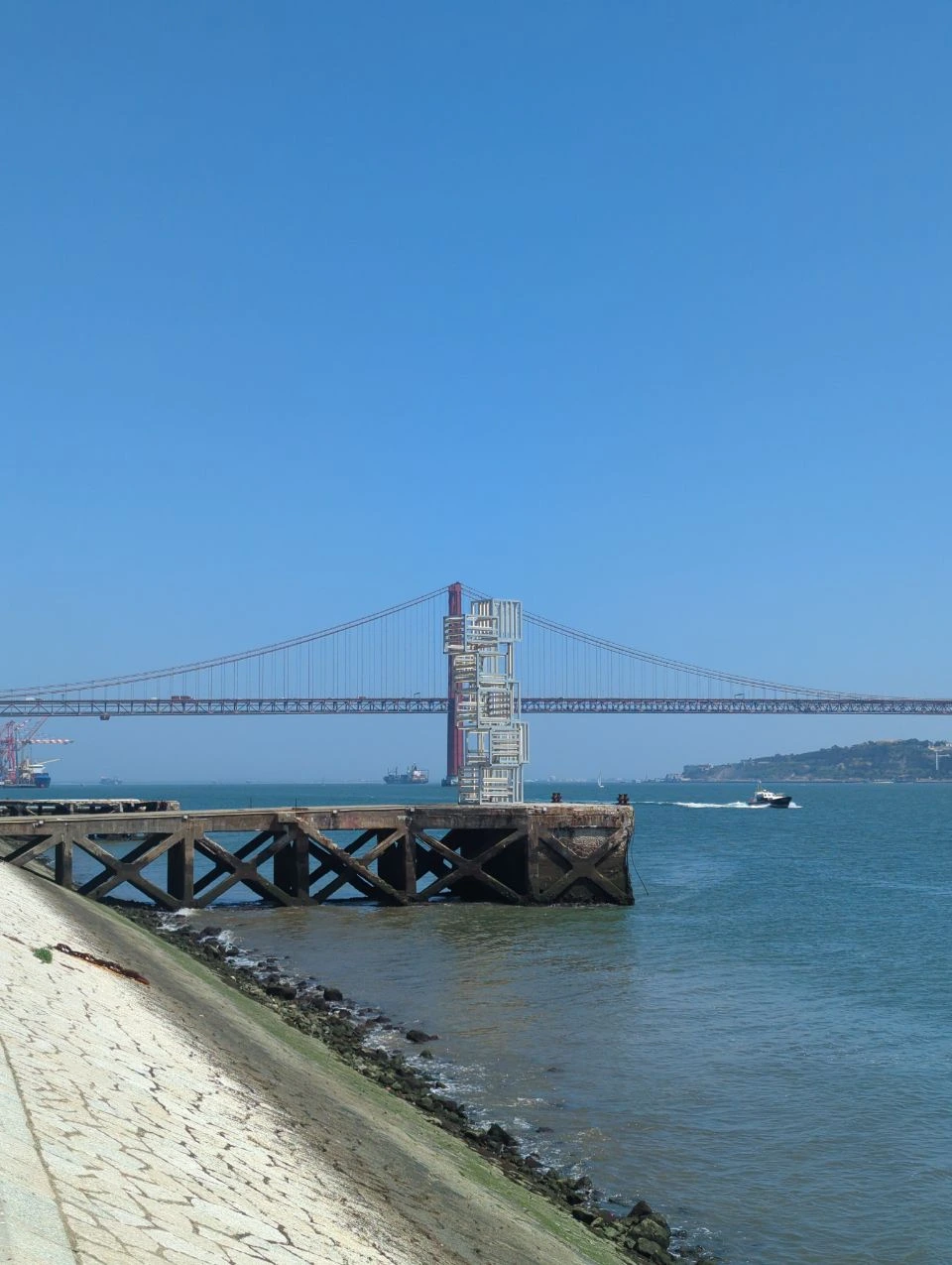 Geometric waterfront artwork frames the 25 de Abril Bridge beside MAAT, with a small boat crossing the Tagus in Lisbon.