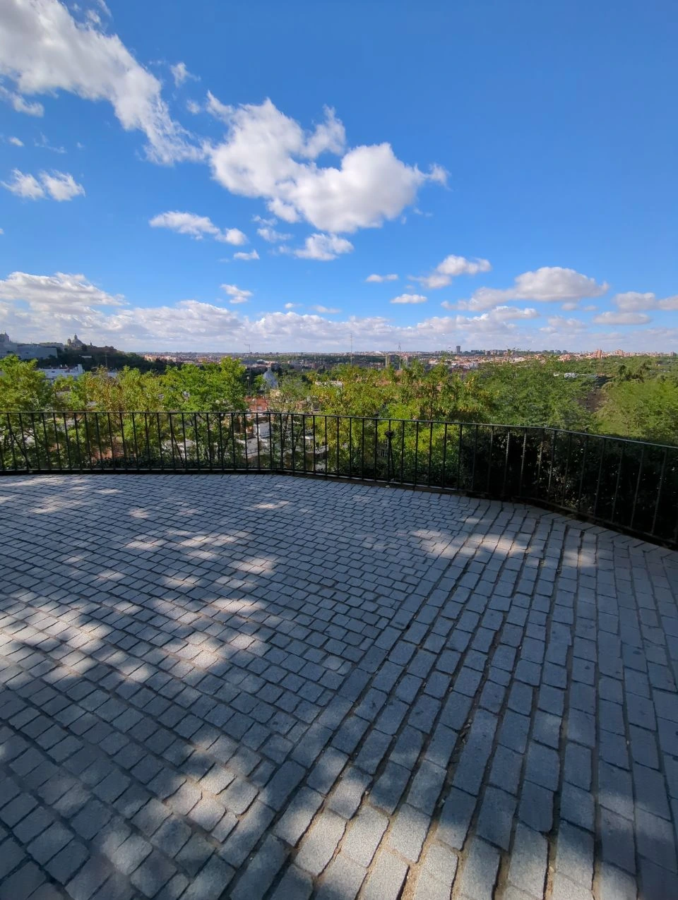 A curved cobbled viewpoint overlooks trees and the Madrid skyline, showing an open proposal setting beneath scattered clouds.