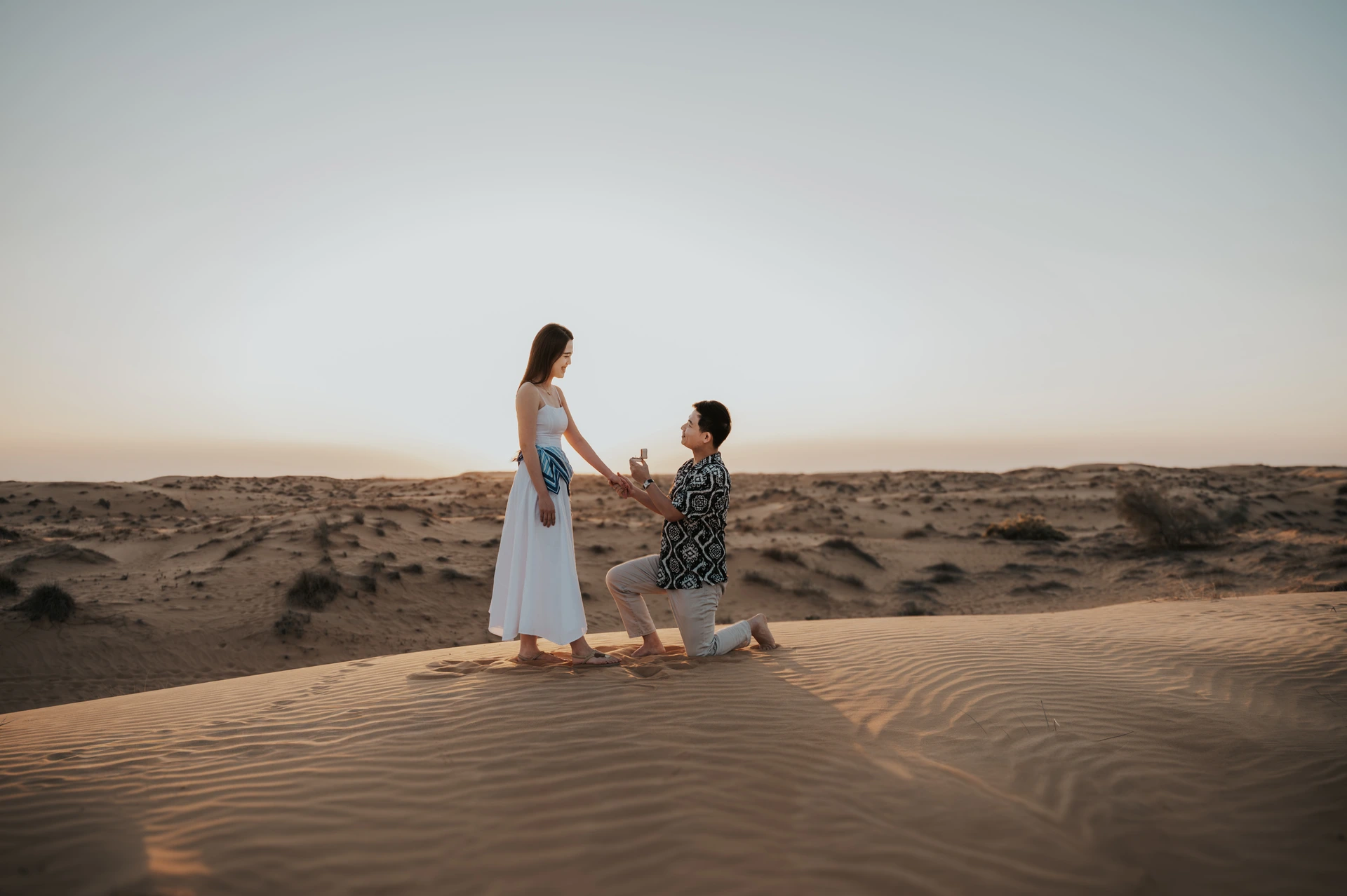 A man kneels on Dubai desert dunes at sunset while his partner reacts, captured during a romantic proposal photoshoot.