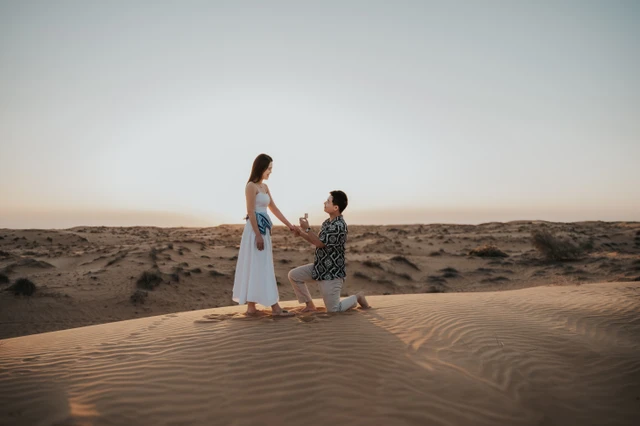 A man kneels on Dubai desert dunes at sunset while his partner reacts, captured during a romantic proposal photoshoot.
