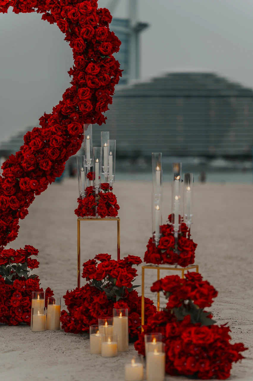 Heart Proposal Setup Dubai - red rose decor by shoreline candles Red rose arrangements and glass candles are styled on Dubai beach beside a heart proposal setup, with Burj Al Arab softly blurred in the distance.