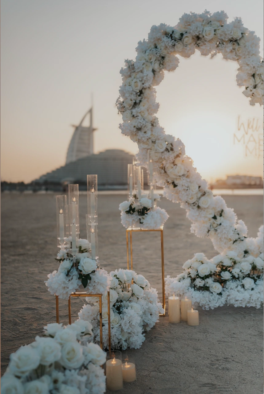 Heart Proposal Setup Dubai - white floral arch decor at dusk White floral arch decor and candles are arranged on Dubai beach at dusk as part of a heart proposal setup with Burj Al Arab behind.
