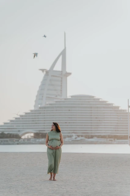 Beach maternity photoshoot Dubai - Burj Al Arab skyline portrait A pregnant woman stands near Dubai shoreline with Burj Al Arab visible behind her during a maternity photoshoot.