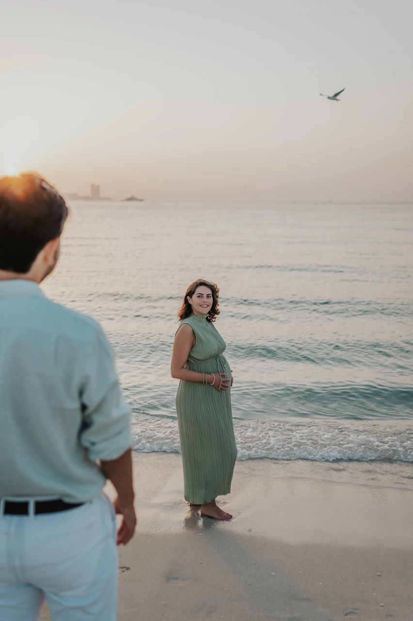 Beach maternity photoshoot Dubai - shoreline bump portrait A pregnant woman in a green dress stands at Dubai shoreline while her partner looks on during a maternity photoshoot.