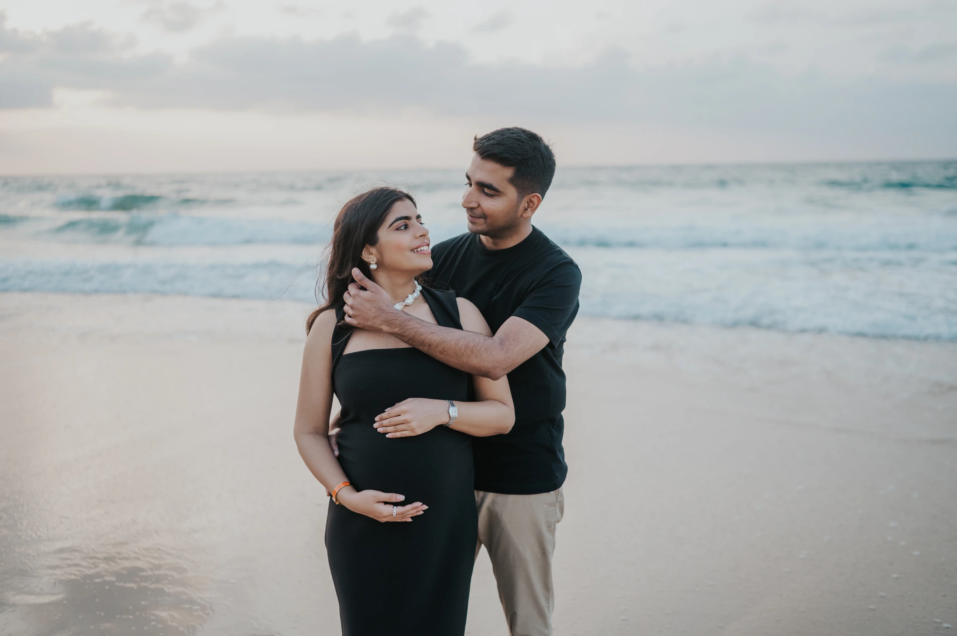 A couple embraces on a calm Dubai beach at dusk during a maternity photoshoot with gentle waves behind them.