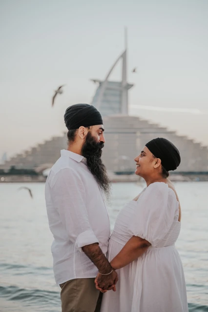 A couple faces each other on Dubai beach at sunset with Burj Al Arab in the background during a maternity photoshoot.