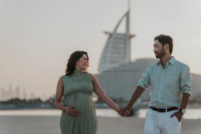 A pregnant woman and her partner hold hands on Dubai beach at sunset with Burj Al Arab behind them.