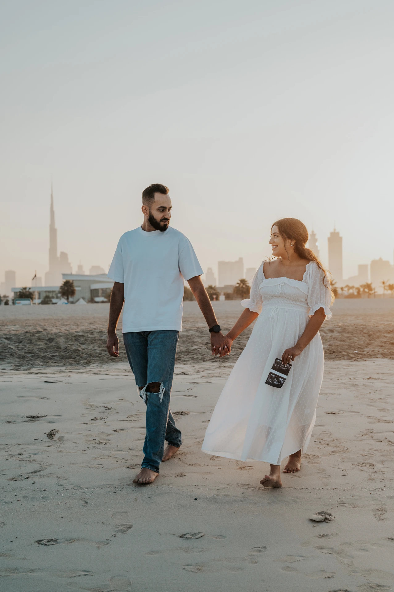 A pregnant couple walks on a Dubai beach with city skyline in the background, combining urban context with a natural maternity photoshoot style.