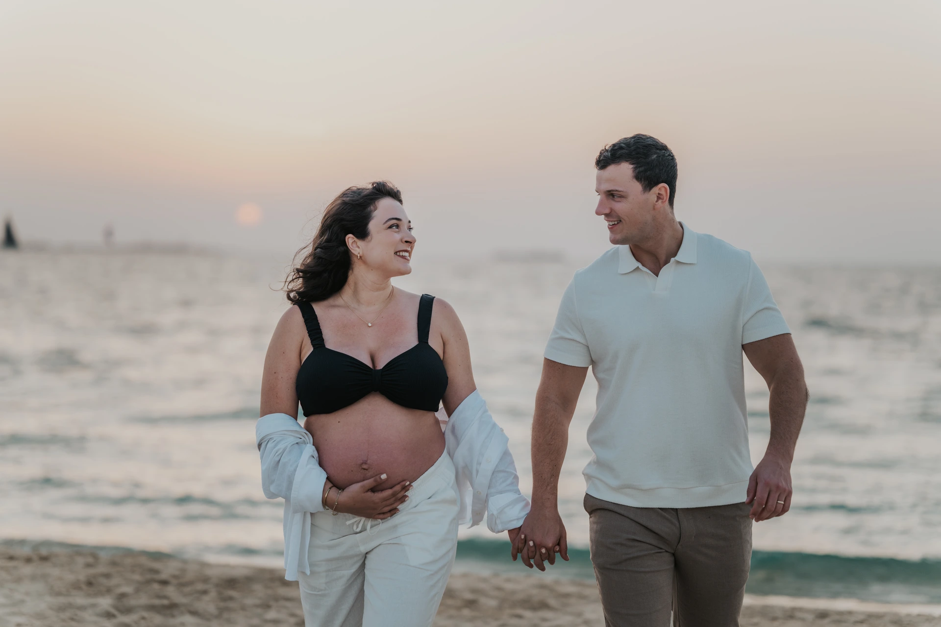 An expecting couple walks by the water at sunset in Dubai during a maternity photoshoot, giving a soft outdoor beach mood with evening light.