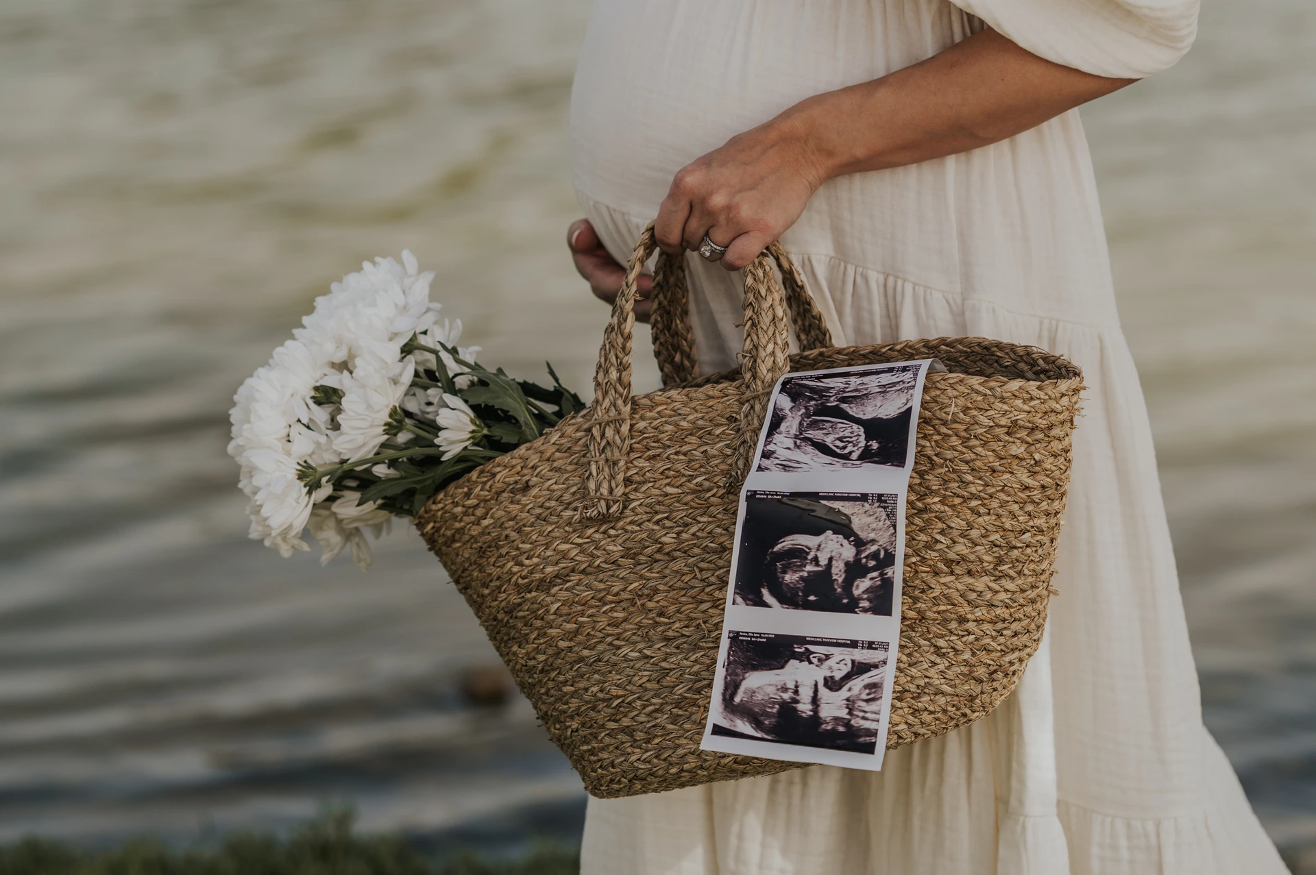 Dubai Maternity Ultrasound Basket by the Water A woven basket holds white flowers and ultrasound images beside a pregnant woman during her Dubai waterfront maternity photoshoot.