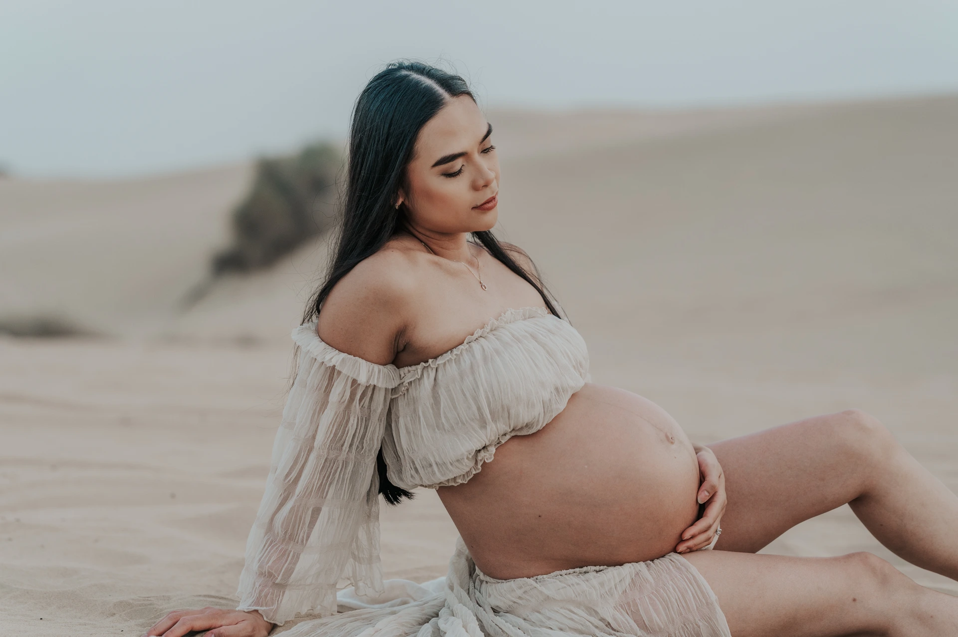 Dubai Desert Maternity Portrait in White A pregnant woman rests one hand on her bump while seated among Dubai desert dunes in a white maternity outfit.