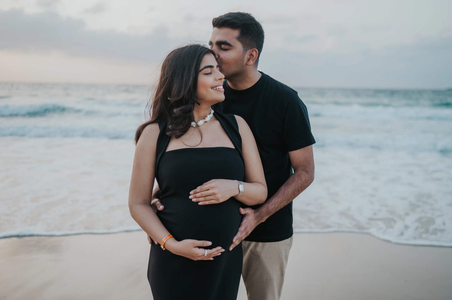 A man kisses his pregnant partner's forehead while they cradle her bump during a Dubai beach maternity photoshoot.
